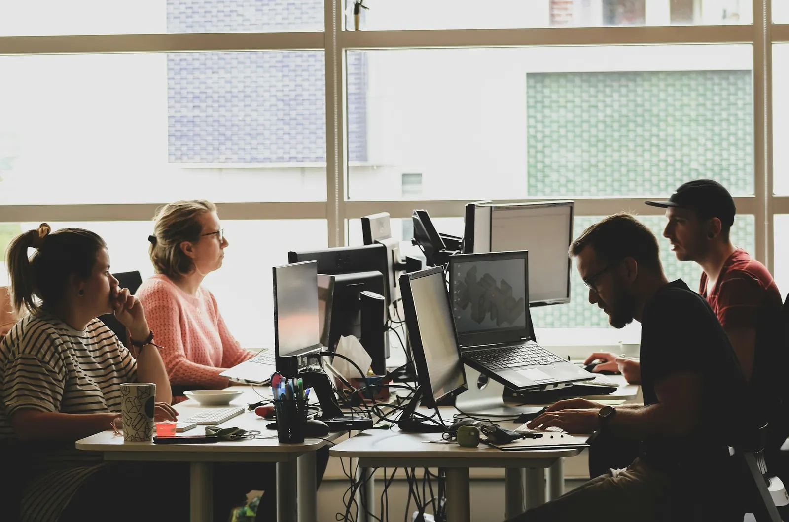 Four people working at computer screens in a bright, sunlit office