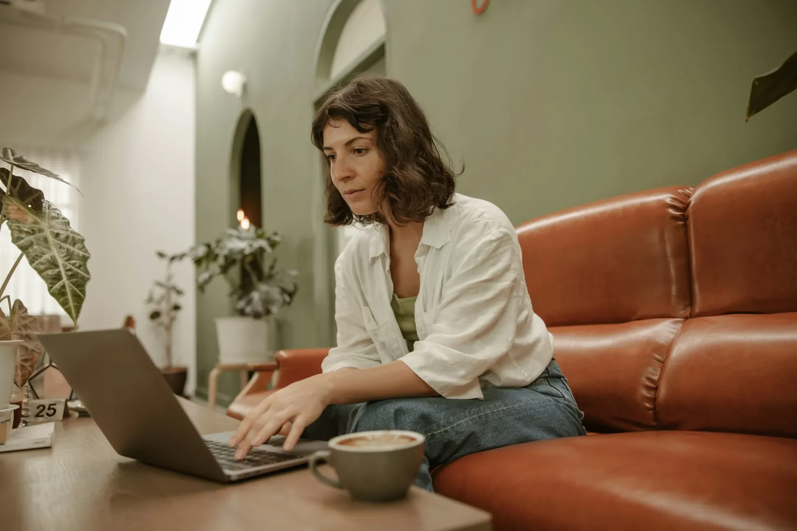 Woman on a leather sofa typing on a laptop, with a cup of coffee on the table